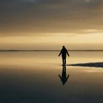 A person standing at water's edge, their reflection showing a freer version of themselves — the visual metaphor for returning to who you were before the world got involved.